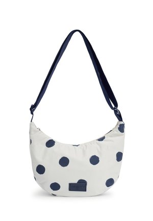 Sac d'épaule blanc en forme de croissant avec une sangle bleu marine et de grands pois bleu marine, doté d'une petite étiquette rectangulaire à l'avant.