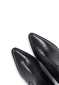 Paire de bottes en cuir noir poli avec des bouts pointus, disposées en diagonale sur un fond blanc.