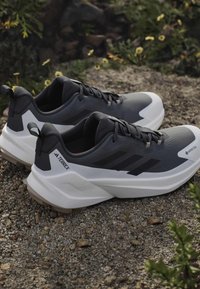 Ζευγάρι από μαύρα και άσπρα παπούτσια Adidas Terrex για τρέξιμο σε μονοπάτι που ξεκουράζονται σε χαλίκι με πράσινο φύλλωμα στο παρασκήνιο.
