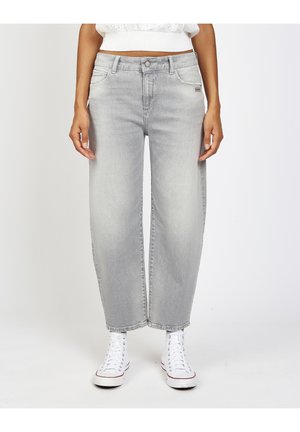 Person trägt hellgraue Jeans mit weitem Bein und weiße High-Top-Sneaker und steht vor einem schlichten weißen Hintergrund.