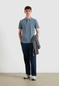 Lichtblauw polo-shirt met knopen, gecombineerd met donkerblauwe broek en een grijze trui over één arm. Model draagt zilveren sneakers.