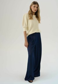 Pull créme, texturé avec des manches ballons associé à un pantalon large marine ; le modèle se tient debout, mettant en valeur la coupe décontractée et le tissu lisse.