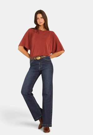 Femme portant un haut ample à manches courtes de couleur rouille, un jean bleu foncé taille haute avec ceinture marron et des bottes marron, debout les mains sur les hanches.