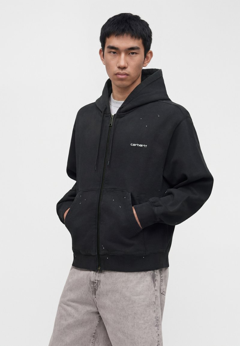 Jeune homme portant un sweat à capuche noir zippé avec un logo blanc et des éclaboussures de peinture, les mains dans les poches, debout devant un fond clair uni.