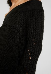 Pull noir à épaules dénudées en maille avec motifs texturés, matière épaisse, encolure large et détails de couture visibles le long des manches.