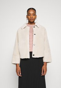 Cream-colored faux fur jacket with a collar, three-button front, and wide sleeves, worn over a pink knitted top and black pleated skirt.