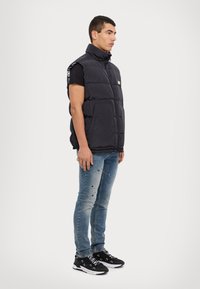 Gilet noir rembourré avec un col haut, sans manches et poches avant. Porté sur une chemise noire et associé à un jean bleu déchiré et des baskets.