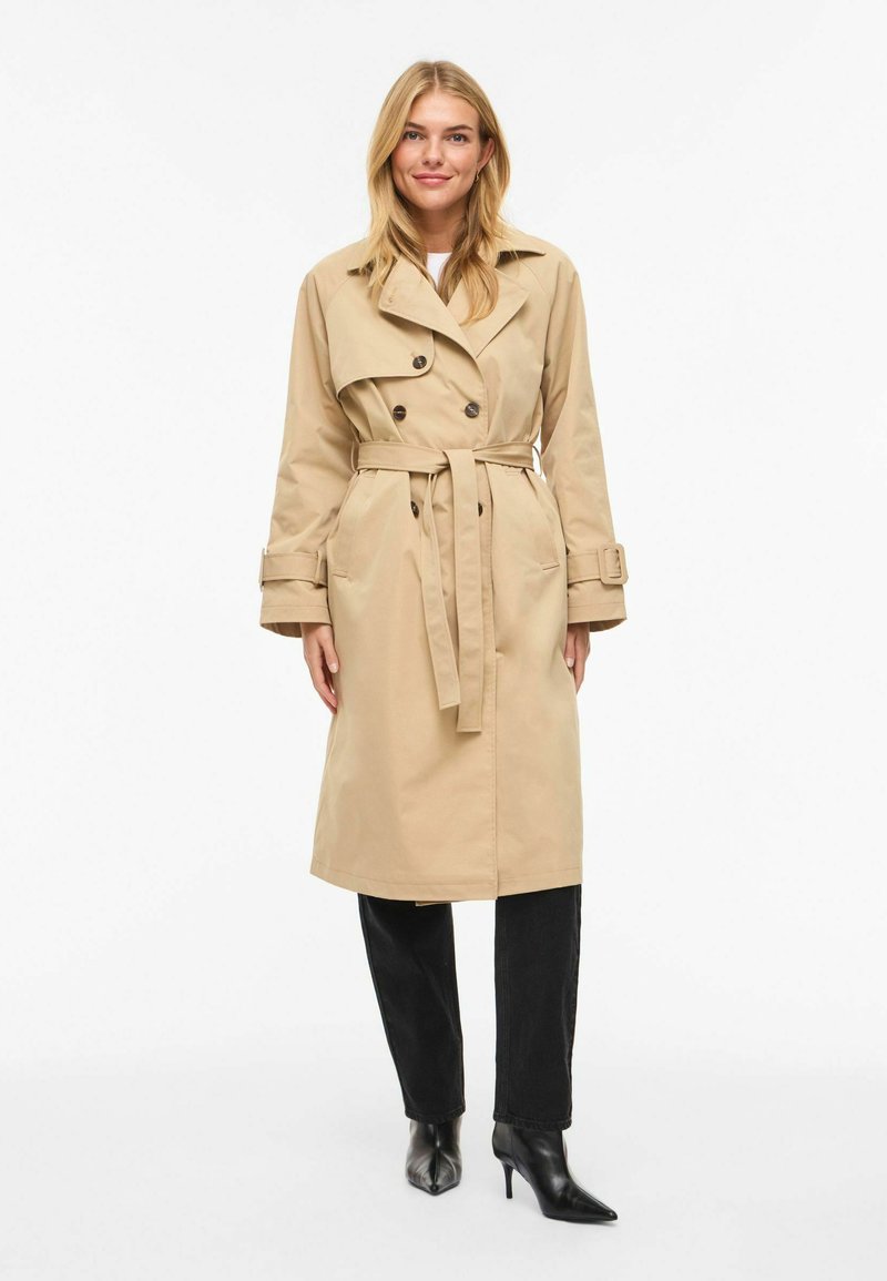 Trench-coat beige avec des boutons double boutonnage, une taille ceinturée et de larges revers, associé à un jean noir et des bottes noires à bouts pointus.