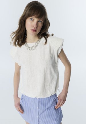 Jeune femme aux cheveux bruns mi-longs, portant un haut blanc texturé et une jupe bleue rayée, posant devant un fond clair.