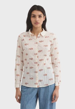 Femme portant une chemise blanche à boutons avec des motifs de poissons rouges et un jean bleu, debout devant un fond uni gris clair.