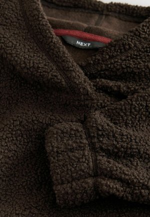 Brunt fleece-stof med en blød, struktureret overflade. Har en krave og en rød detalje på det indre label. Varm og hyggelig udseende.