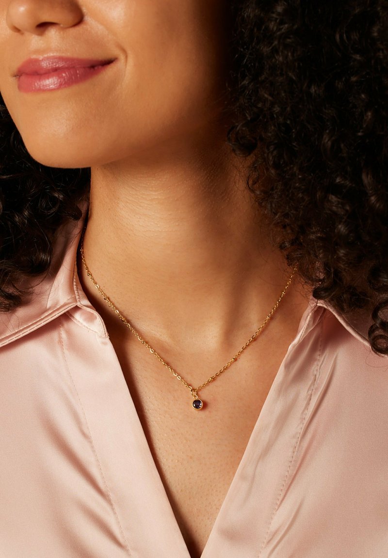 Gouden ketting met een kleine ronde paarse hanger, gedragen over een lichtroze satijnen blouse. Eenvoudig ontwerp met een glanzende afwerking.