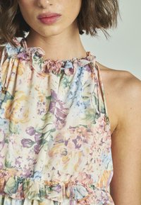 Top in tessuto leggero con motivo floreale, caratterizzato da scollatura e orlo arricciati, con colori pastello che includono accenti di rosa, blu e giallo.