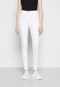 Jeans skinny brancos de cintura alta com uma textura suave, apresentando cinco bolsos e um fecho frontal com botão, combinados com ténis claros.