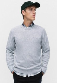 Grå stickad tröja med rundad halsringning, ribbade ärmslut och avslut, lagd över en blå och vit rutig skjorta, tillsammans med en mörk keps.