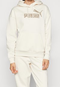 Kremowa bluza z kapturem, wykonana z miękkiego materiału, z przednią kieszenią, kapturem ze sznurkiem i logo Puma z wzorowanym designem.