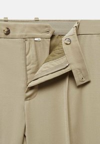 Pantalon beige avec fermeture éclair et braguette à boutons partiellement ouverte, montrant la doublure intérieure et les boutonnières sur la ceinture et le rabat de la braguette.