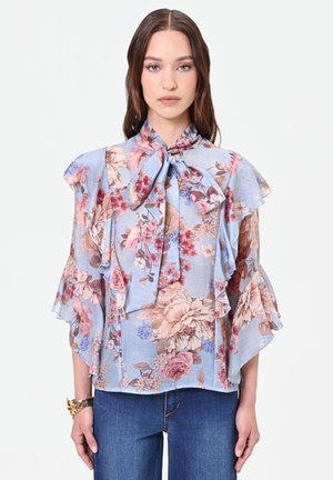 Donna che indossa una camicetta azzurro chiaro a fiori con un grande fiocco al collo e maniche arricciate, abbinata a jeans blu.