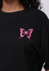 Svart bomull T-skjorte med et rosa sommerfugl-trykk på venstre brystområde. Designet er detaljert med vingemønstre og teksturer.