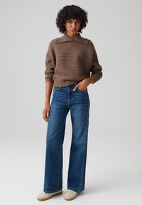 Maglione marrone a coste con colletto. Abbinato a jeans in denim blu a gamba larga. Sneakers beige con leggeri accenti e suole in gomma.