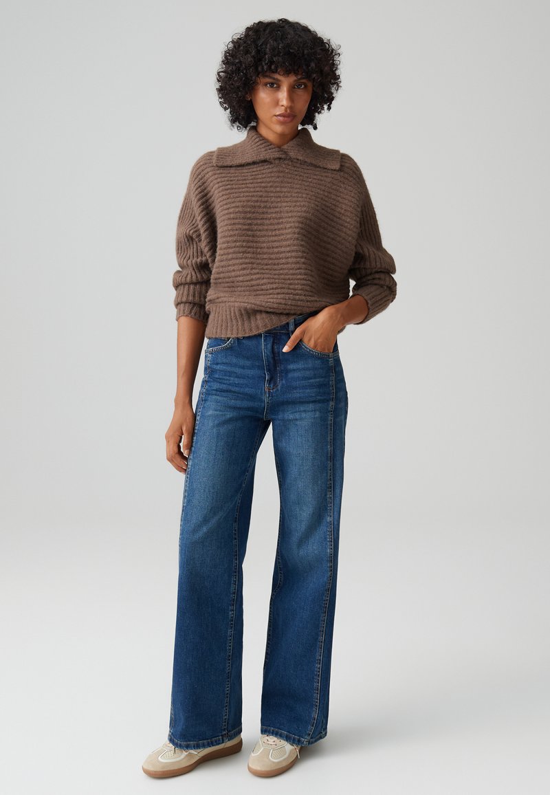 Maglione marrone a coste con colletto. Abbinato a jeans in denim blu a gamba larga. Sneakers beige con leggeri accenti e suole in gomma.