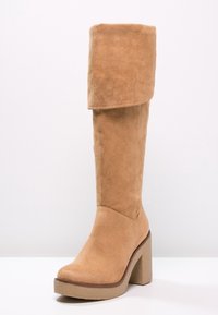 Botte haute jusqu'au genou en daim beige avec revers plié, talon haut épais et semelle plate-forme sur fond blanc uni.