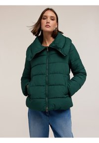 Tmavě zelená péřová bunda s širokým límcem, horizontálním prošíváním, zapínáním na zip a bočními kapsami. V kombinaci s krásně modrými džíny.