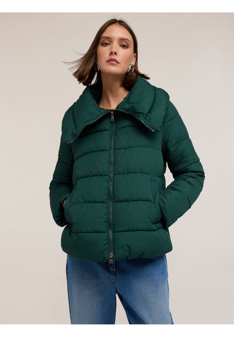 Tmavě zelená péřová bunda s širokým límcem, horizontálním prošíváním, zapínáním na zip a bočními kapsami. V kombinaci s krásně modrými džíny.