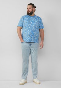 Blaues T-Shirt mit kurzen Ärmeln und weißem Blättermuster, kombiniert mit hellblauen Jeans und weißen Sneakers. Vor einem neutralen Hintergrund.