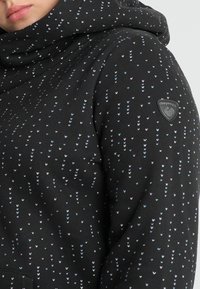Primo piano di una felpa nera con piccoli motivi a forma di cuori blu e rosa e una patch del logo del brand sulla parte superiore del braccio.