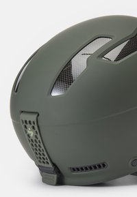 Zielony, matowy kask narciarski z akcentami z włókna węglowego, aerodynamiczny kształt, grille wentylacyjne oraz tylna, regulowana taśma z heksagonalnym wzorem.