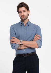 Camisa de botones azul claro con mangas largas, arremangadas en los puños, combinada con pantalones de cuadros oscuros y un cinturón de cuero marrón. Reloj plateado en la muñeca.