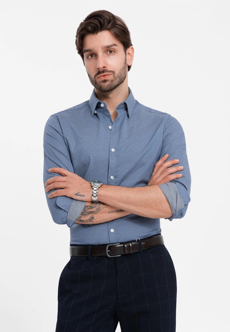 Camisa de botones azul claro con mangas largas, arremangadas en los puños, combinada con pantalones de cuadros oscuros y un cinturón de cuero marrón. Reloj plateado en la muñeca.