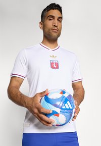 Camisa atlética blanca con detalles rojos y un logo dorado, combinada con un balón de fútbol multicolor que presenta remolinos azules, rojos y blancos.