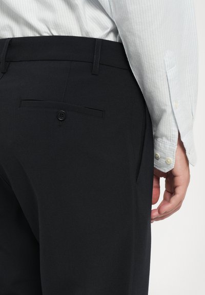 Dunkle Hosen aus glattem Stoff, mit einer Gesäßtasche mit Knopfverschluss und sichtbaren Nahtdetails. Das Hemd hat ein leicht gestreiftes Muster.
