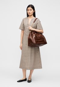 Καφέ δερμάτινη τσάντα tote με χαλαρή σχεδίαση και χρυσή αλυσίδα ως διακόσμηση, δίπλα σε μπεζ φόρεμα μέχρι το γόνατο με καρό μοτίβο. Μαύρα επίπεδα παπούτσια.