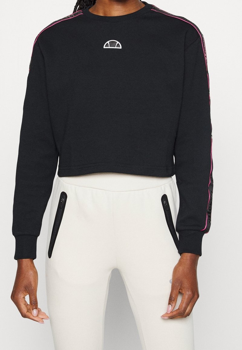 Zwarte cropped sweatshirt met een cirkelvormig logo, roze zijstrepen en geribbelde manchetten, gecombineerd met lichtgekleurde sweatpants met ritszakken.