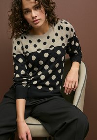 Sweter w czarne i beżowe kropki, wykonany z dzianiny, z okrągłym dekoltem, długimi rękawami i ściągaczami. Stylizowany z czarnymi spodniami.