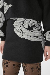Zwarte gebreide trui met een grijs rozenpatroon op de mouwen en taille, voorzien van een zachte textuur en een aansluitend ontwerp. Zwarte panty's met bloemendessin.