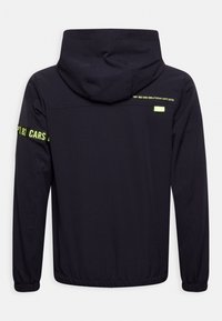 Dark navy jacket with a hood, featuring elastic cuffs, a back logo in reflective yellow, and a smooth, lightweight fabric texture.