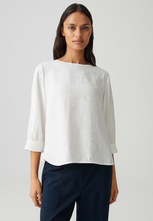 Vrouw met lang donker haar, gekleed in een witte blouse met lange mouwen en donkerblauwe broek, staand tegen een effen lichte achtergrond.