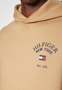 Felpa con cappuccio color camoscio con coulisse. Presenta la scritta "HILFIGER NEW YORK" in caratteri scuri e un piccolo logo a bandiera in rosso, bianco e blu sotto la scritta "Est. 1985."