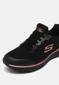 Melnas sporta kurpes ar teksturētu sietu augšdaļu, sintētiskām akcentiem, auklu dizainu un rozā zelta svītru gar zoli.