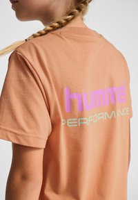Ferskenfarvet bomulds t-shirt med korte ærmer, prydet med et iøjnefaldende lilla "hummel" logo og lyseblå "PERFORMANCE" tekst på ryggen.