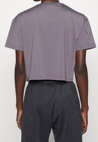 Person som bär en lös, kort lila t-shirt och mörkgrå byxor med elastisk midja, sedd bakifrån.