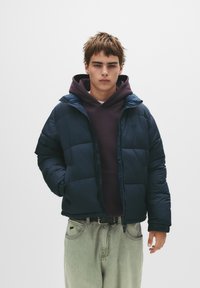 Jachetă pufosă navy cu glugă, având un design matlasat, purtată peste un hanorac mov închis, asortată cu blugi largi de culoare deschisă.