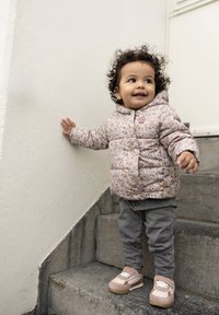Ljusrosa puffjacka med blommönster, grå jeans och beige skor. Barnet står på en trappa och stödjer sig mot väggen.