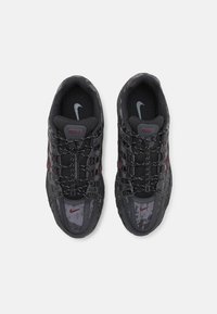 Sapatilhas desportivas pretas com um exterior texturizado, apresentando detalhes em vermelho e um sistema de atacadores único. Logótipo da Nike na lingueta e na lateral.