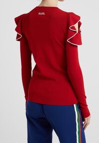 Top rosso a maniche lunghe con maniche arricciate e spalle scannellate, con bordi bianchi. Logo "KARL" posizionato sul retro del collo.