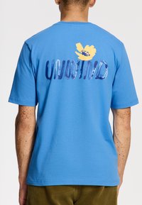 Camiseta de algodón azul con mangas cortas. Presenta un gráfico de una flor amarilla y la palabra "DESCONECTAR" en texto azul en la parte de atrás. Textura suave.